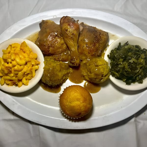 Soul food plate with gravy-covered chicken, mac and cheese, collard greens, cornbread dressing, and a muffin.